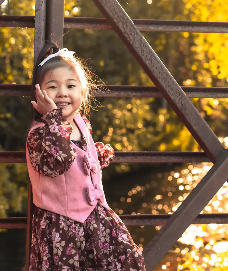 A little girl in a floral dress smiles and waves from a bridge, backlit by glowing autumn leaves