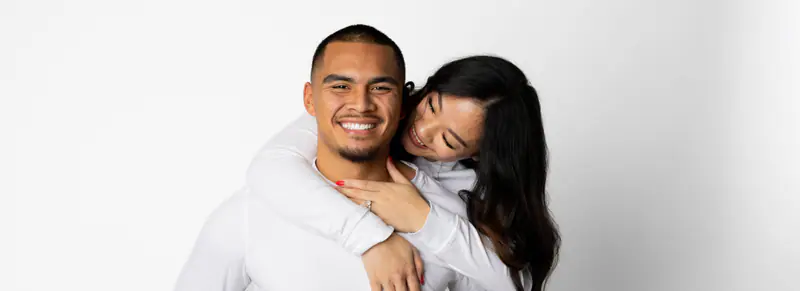 A laughing couple in white embrace in the studio, showing an engagement ring
