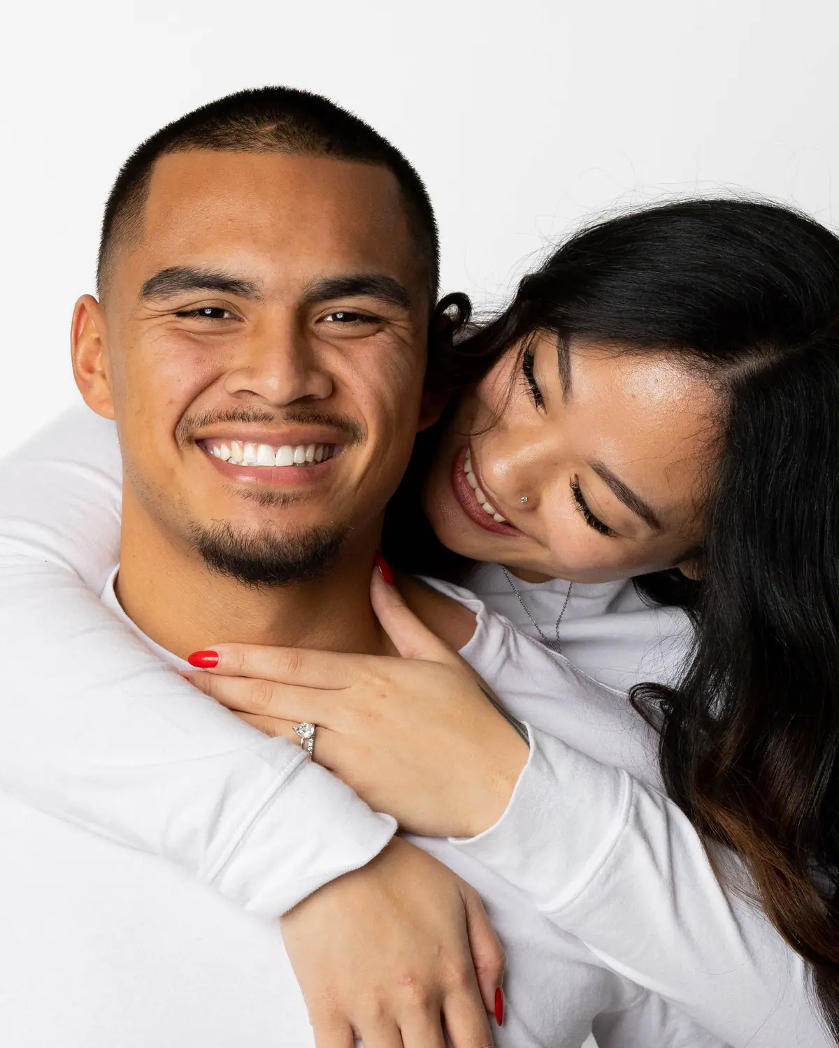 A laughing couple in white embrace in the studio, showing an engagement ring