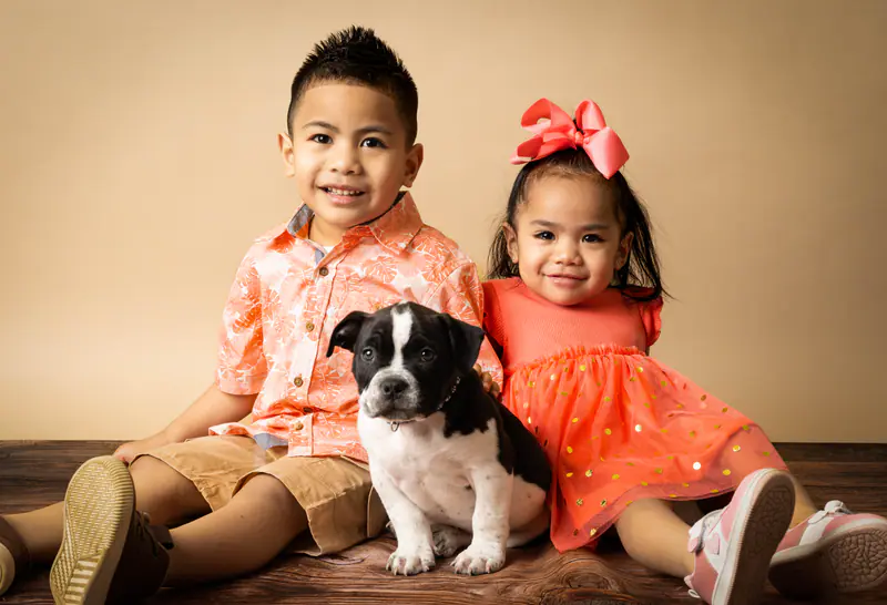 A brother and sister in coral outfits sit with their black and white puppy in the studio