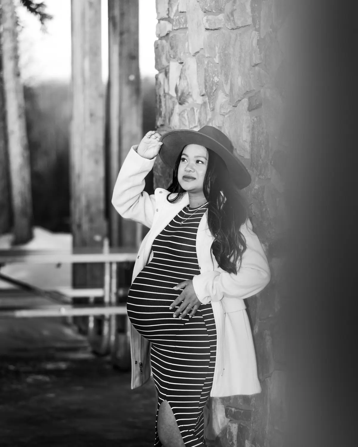 Black and white maternity portrait of a mother in a felt hat and striped dress beside a stone pillar