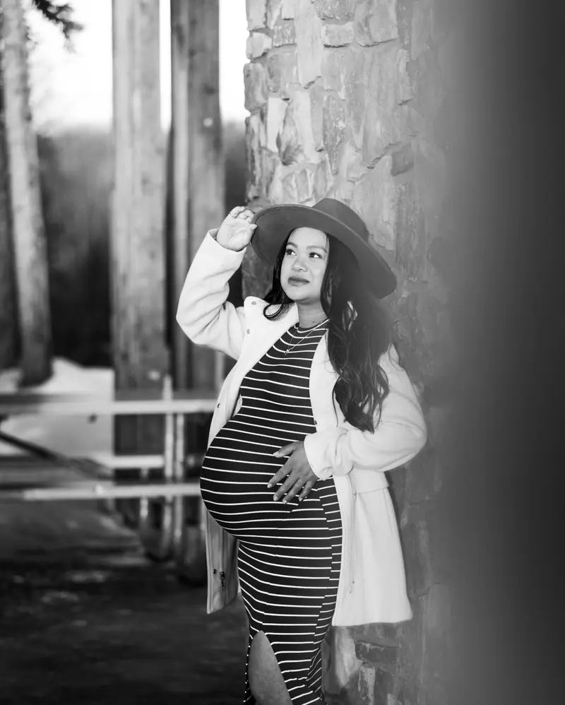Black and white maternity portrait of a mother in a felt hat and striped dress, cradling her bump beside a stone pillar