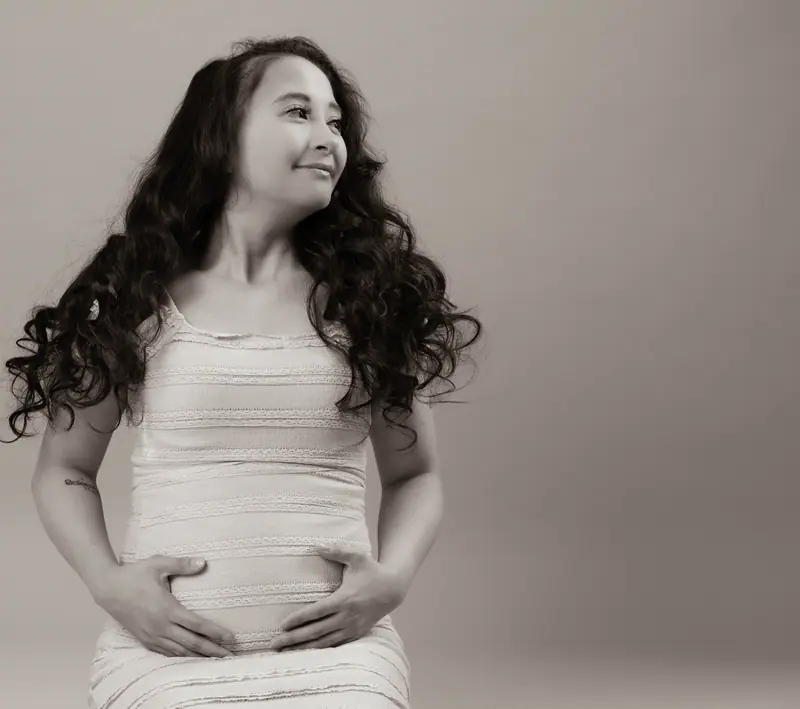 Sepia studio maternity portrait of a mother with long curly hair, hands resting on her bump