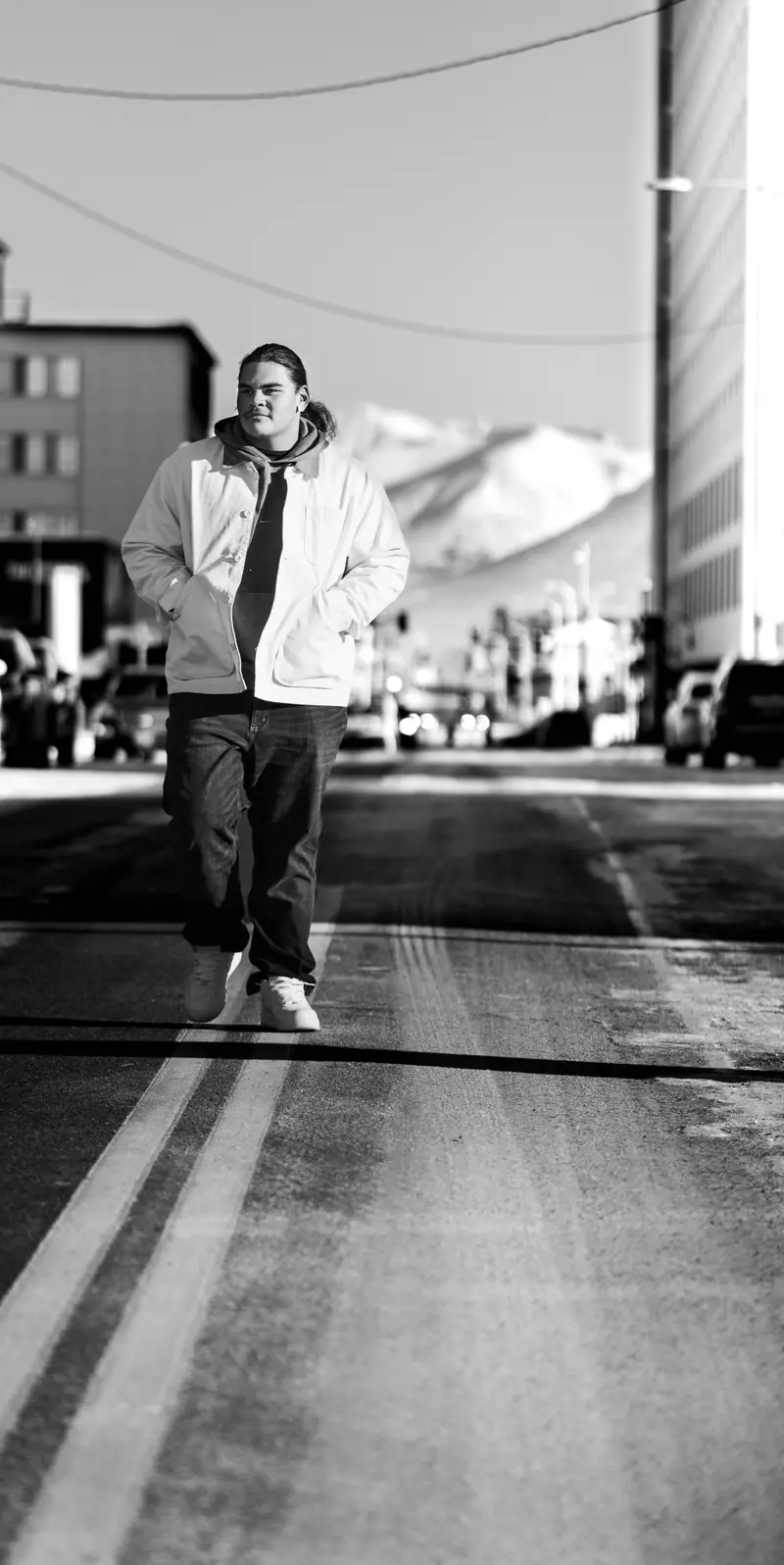 Black and white senior portrait of a young man walking down a snowy downtown Anchorage street with the Chugach Mountains behind