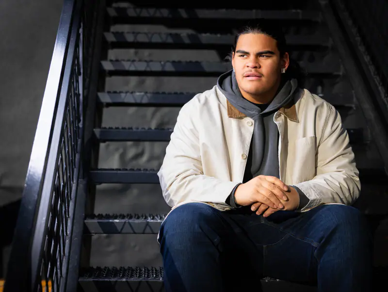 A senior in a cream chore coat sits on dark metal stairs in warm window light