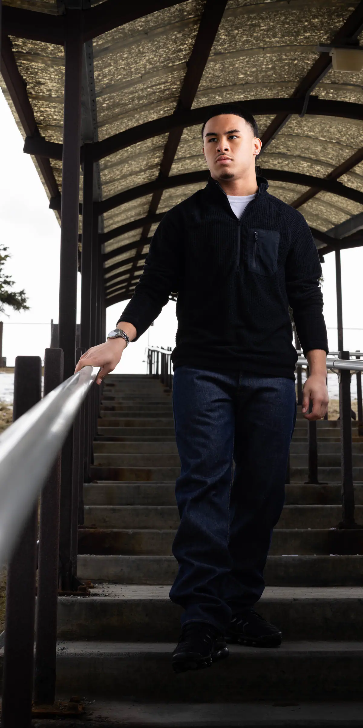 A young man in a black pullover walks down covered outdoor stairs, one hand on the rail