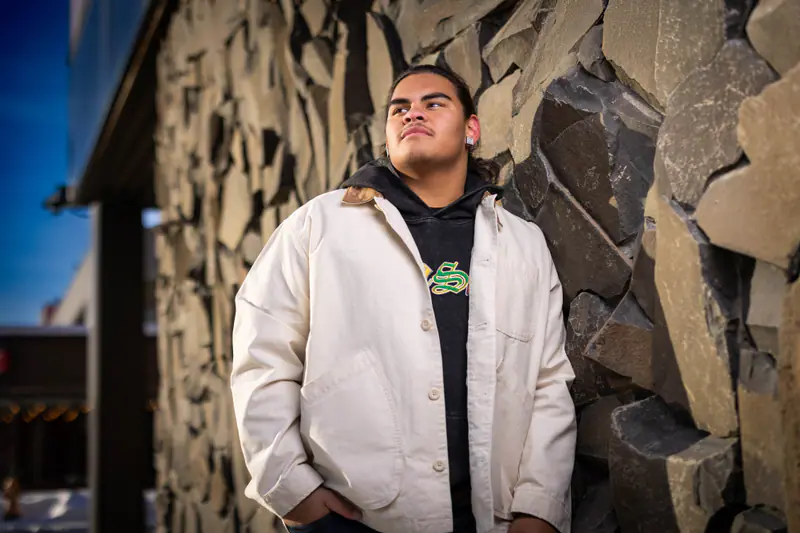 A senior in a cream jacket leans against a slate stone wall under a bright blue sky